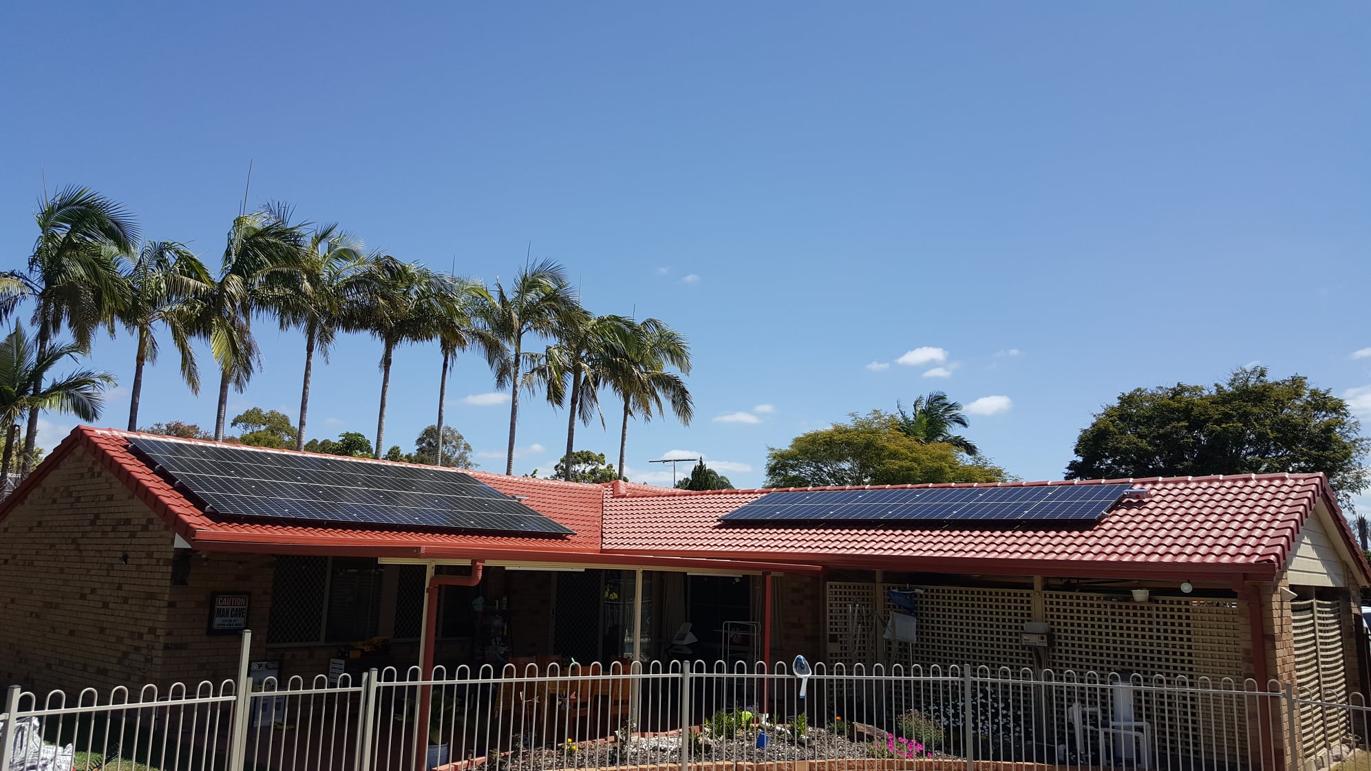 Single storey house with red roof and solar panels, palm trees in background with blue skies